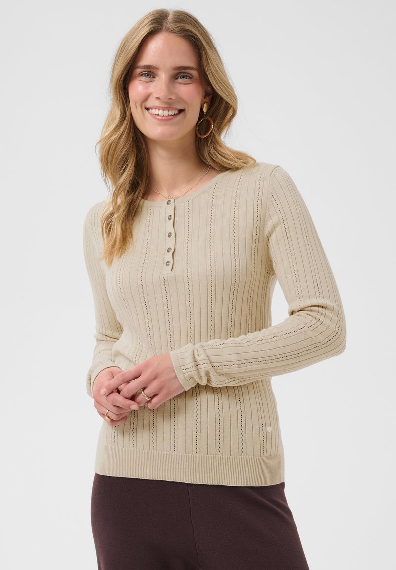 Femme souriante, portant un haut beige en maille côtelée à manches longues avec boutons, des créoles et un pantalon foncé, sur un fond uni clair.