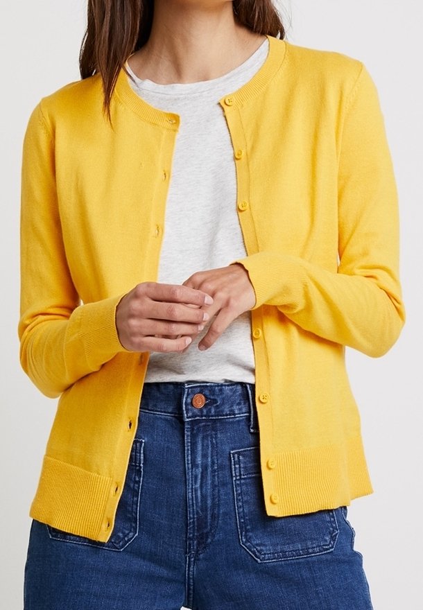 Gul kofta gjord av mjukt stickat material, har en rund halsringning, knappstängning och ribbade ärmslut och nederkant. Bärs över en grå topp.