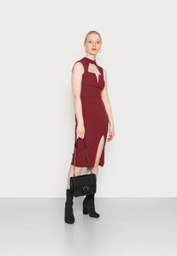 Vestido midi burgundy com decote alto e detalhes recortados, apresentando uma fenda lateral. Combinado com uma carteira preta e botas de tornozelo.