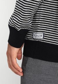 Maglione a righe bianche e nere con una texture a coste. Presenta un'etichetta logo e polsini aderenti. Abbinato a pantaloni grigi.