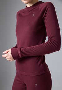 Burgundská športová blúzka s dlhým rukávom a vysokým golierom, vyrobená z jemného materiálu. Obsahuje otvory na palce a malé logo nad lemom.