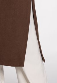 Brun, tekstureret stofjakke med skræddersyet design og sideslids, kombineret med cremefarvede bukser, der viser en kontrasterende farvepalette.