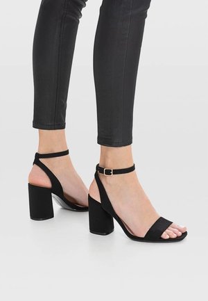 Sandales à talons noires avec un design à orteil carré, en suede. Équipées d'une fine bride de cheville et d'un talon cubique pour plus de stabilité.