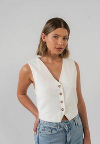 Weiße ärmellose Bluse mit tiefem V-Ausschnitt und fünf goldenen Knöpfen auf der Vorderseite. Aus strukturiertem Stoff gefertigt, mit einem cropped Design.