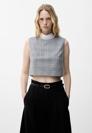 Jonge vrouw met blond haar draagt een grijs geruit cropped top zonder mouwen over een witte coltrui en zwarte broek met riem.