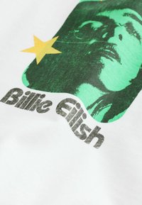 White t-shirt featuring a green graphic of a face and a yellow star. Text below reads "Billie Eilish" in bold black lettering.