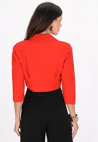 Blazer rosso tagliato con maniche a tre quarti, spalle sagomate e tessuto liscio. Pantaloni neri a vita alta e bracciale dorato al polso.