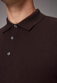 Polo de punto marrón con una textura suave, que presenta un cuello y tres botones grises en la parte delantera.