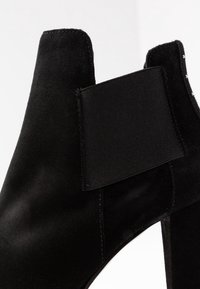 Černé semišové kotníkové boty s elastickou boční částí, hladkým povrchem, zaoblenou špičkou a vysokým podpatkem. Bez viditelného kování nebo vzorů.