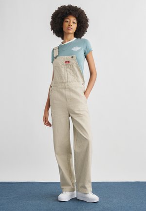 Jonge vrouw met afrohaar, gekleed in beige gestreepte tuinbroek, blauw t-shirt en witte sneakers, staand op een blauw tapijt tegen een witte achtergrond.