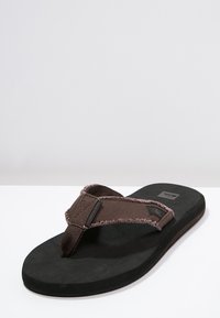 Barna flip-flop szandál, textúrált szövetpánttal, puha fekete talppal és címke részlettel a pánton. Laposan megtervezett, egyszerű dizájn, amely alkalmas hétköznapi viseletre.