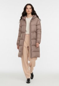 Jasna brązowa puchowa kurtka z kapturem, wykonana z pikowanego materiału. Posiada zamek z przodu, dwie boczne kieszenie oraz długi, luźny krój.