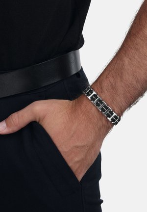 Braccio maschile con un bracciale in metallo nero e argento, mano in tasca dei pantaloni scuri, indossando una camicia nera con una cintura in pelle nera.