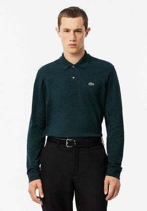 Polo a maniche lunghe verde con colletto e un piccolo logo di coccodrillo verde sul lato sinistro del petto, abbinato a pantaloni neri e una cintura.