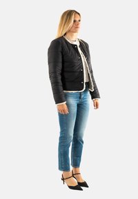 Chaqueta negra tipo puffer con un acabado texturizado, cierre de botonadura al frente y ribete de pelo en el cuello, llevada sobre una blusa blanca y combinada con jeans azules y tacones negros.