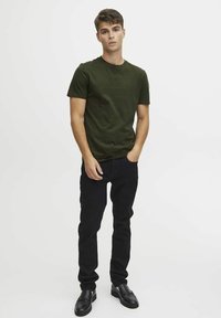 Olivengrønn kortermet t-skjorte, slim-fit. Kombineres med svarte jeans og svarte loafers. Modellen står med hendene i lommene, nøytral bakgrunn.