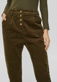Pantalon en velours côtelé marron taille haute avec quatre boutons à l'avant et poches latérales, porté avec un haut à manches longues côtelé vert olive.