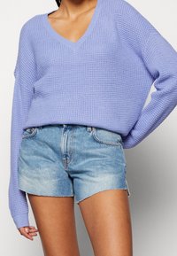 Jasnofioletowy sweter dzianinowy z dekoltem w serek i oversize'owymi rękawami, zestawiony z postrzępionymi szortami dżinsowymi z wystrzępionymi krawędziami.