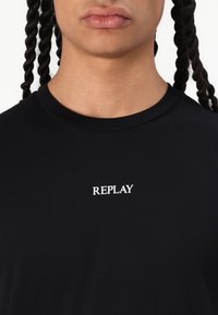 černé tričko s kulatým výstřihem, na hrudi s bílým textem "REPLAY". Vyrobeno z hladké, měkké látky s přiléhavým střihem.