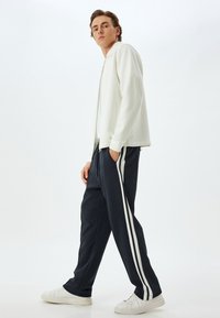 Hvid zip-up jakke, navy sweatpants med hvide sidelister og hvide sneakers. Model står oprejst mod en ensfarvet baggrund.