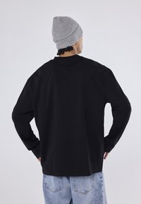 Cămașă neagră cu mâneci lungi și guler rotund, asociată cu blugi din denim de un albastru deschis și o căciulă gri cu nervuri. Material neted cu o croială lejeră.