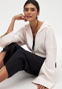 Lichtbeige oversized shirt met brede mouwen, gedragen over een zwarte top en broek. Lange zwarte ketting met een ronde hanger als accent.