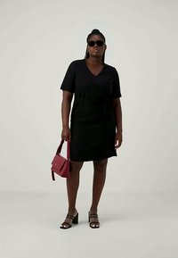 Črna kratka oblekica s kratkimi rokavi in V-izrezom ter zategnjenim pasom. Poudarjena z črnimi blok petami in rdečo torbico čez ramo. Gladka tekstura tkanine.