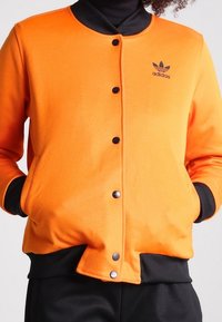 Orange sweatshirtjacka med svarta detaljer, tryckknappar och en Adidas-logotyp. Tillverkad av mjukt tyg med sidofickor.