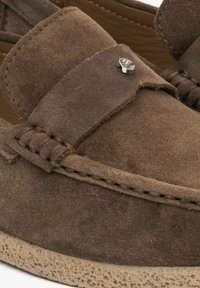 Bruine suede instappers met een gestikt ontwerp en een kleine metalen accent. De zool is voorzien van een textuur in een lichte beige kleur.