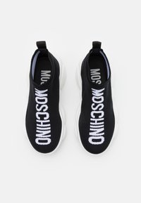 Černé slip-on tenisky vyrobené z texturovaného pleteného materiálu s výrazným bílým nápisem "MOSCHINO" a bílými gumovými podrážkami. Minimalistický design s poutkem.