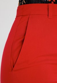 Detailní záběr na červené kalhoty s vysokým pasem, boční kapsou a poutky na opasek, které jsou doplněny černým květinovým topem.