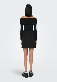 Svart avlang topp uten skuldre, tettsittende lange ermer, kombinert med et kort svart semsket skinnskjørt. Modellen har på seg kraftige svarte sko med hvite sokker.