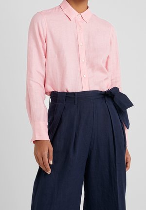 Personne portant une chemise rose clair à manches longues boutonnée, rentrée dans un pantalon taille haute bleu marine avec une ceinture nouée sur le côté.