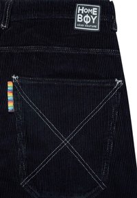 Crne korduroy hlače s bijelom etiketom s natpisom "HOME BOY LOUD COUTURE", višebojnim prugastim akcentom na džepu i kontrastnim bijelim šavovima.
