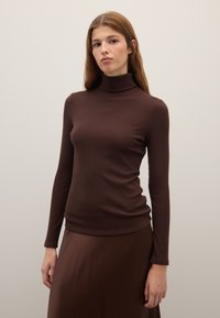 Brown ribbed long-sleeve top with a high neckline. Fabric appears soft, fitted design with a subtle texture. Model pairs it with a matching skirt.