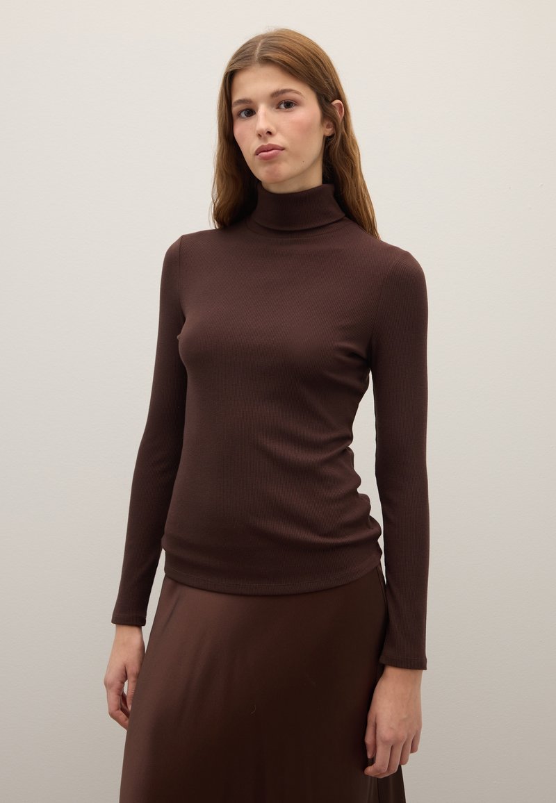 Brown ribbed long-sleeve top with a high neckline. Fabric appears soft, fitted design with a subtle texture. Model pairs it with a matching skirt.