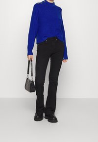 Person klädd i en blå tröja med guldknappar, svarta tajta byxor, svarta kraftiga stövlar, som håller en liten svart handväska.