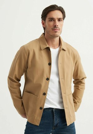 Veste marron clair en coton avec un col de chemise, fermeture à boutons sur le devant et deux poches latérales. Portée par-dessus un t-shirt blanc.
