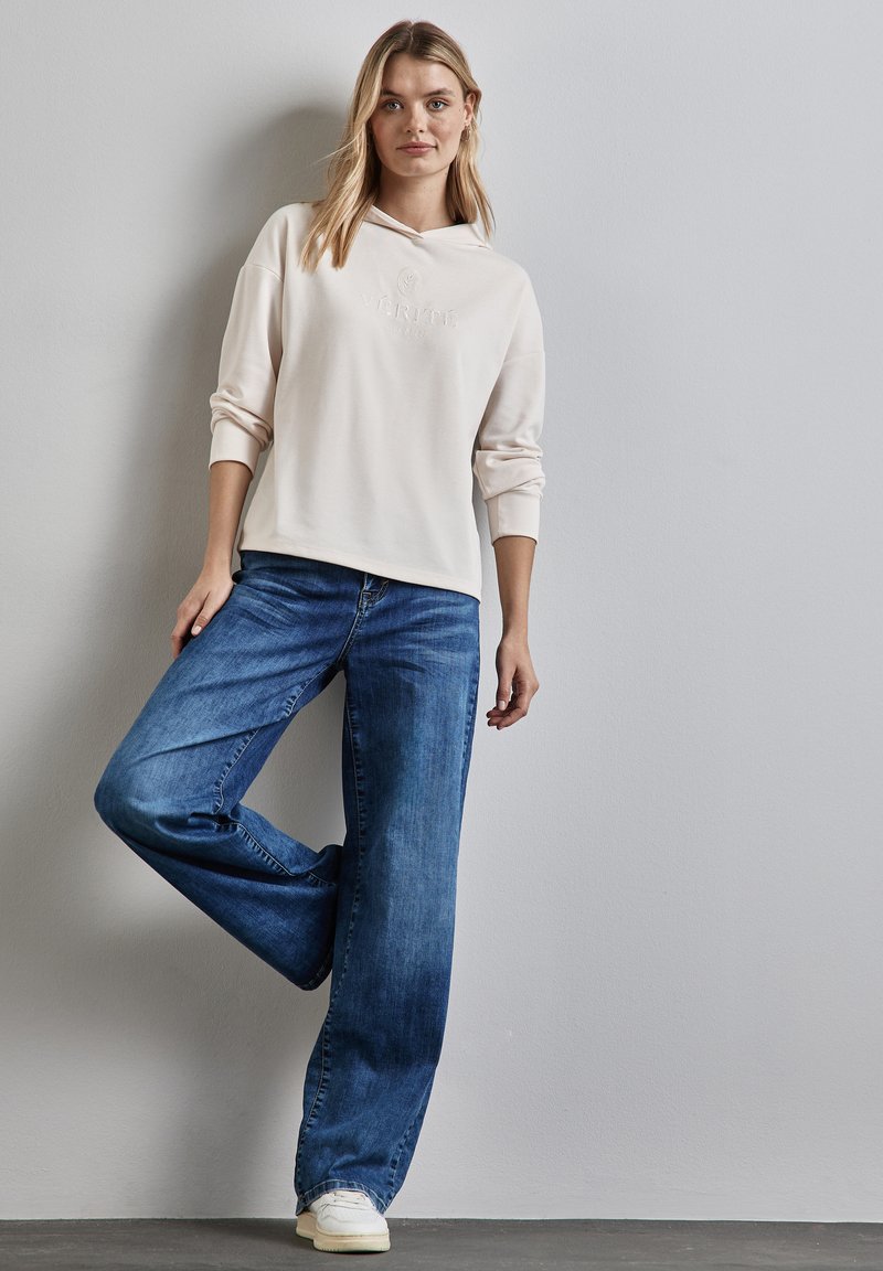 Helle beige Kapuzenjacke mit gesticktem Logo, kombiniert mit weit geschnittenen blauen Jeans und weißen Sneakers, posiert vor einem neutralen Hintergrund.