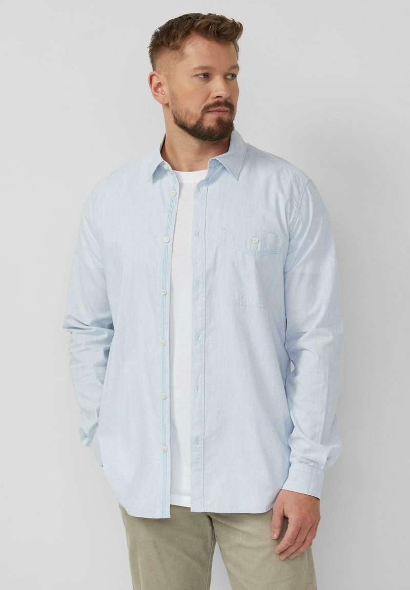 Man met kort bruin haar en baard, gekleed in een lichtblauw overhemd over een wit T-shirt en beige broek, staand tegen een effen achtergrond.