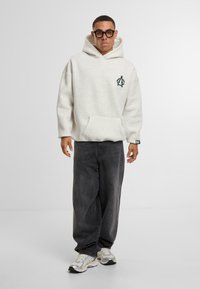 Felpa grigia chiara con cappuccio, tasca frontale e logo verde scuro, abbinata a jeans grigi larghi e sneakers riflettenti.