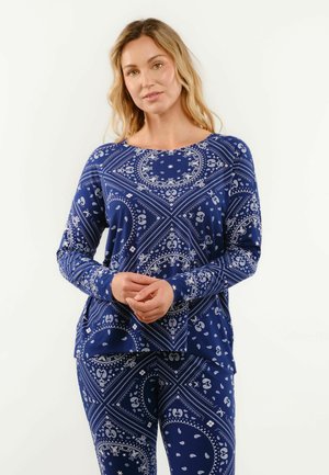 Kobieta o blond włosach, ubrana w pasującą niebieską bluzkę z długim rękawem i spodnie z białymi wzorami paisley i geometrycznymi, stojąca na jednolitym tle.