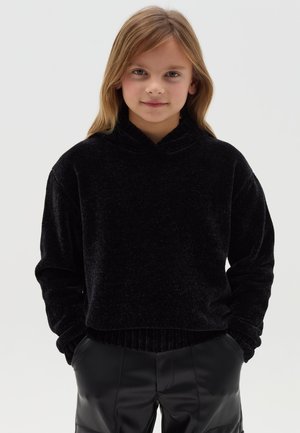 Sudadera de terciopelo negro con cuello alto, mangas largas y dobladillo acanalado. Combinada con pantalones de cuero sintético negro. Textura suave, diseño minimalista.