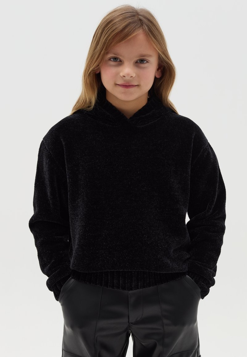 Sudadera de terciopelo negro con cuello alto, mangas largas y dobladillo acanalado. Combinada con pantalones de cuero sintético negro. Textura suave, diseño minimalista.