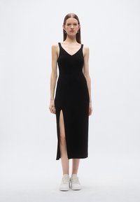 Čierne rebrované midi šaty s výstrihom do V, priliehavým strihom, bočným rozparkom a tenkými ramienkami, v kombinácii s bielymi teniskami.
