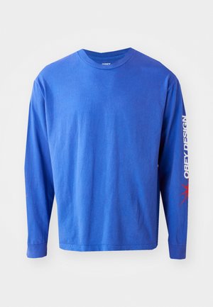 Langærmet skjorte i livlig blå bomuldsstof. Har en rund halsudskæring, trykt tekst "OBEY DESIGN" i hvid langs ærmet og røde detaljer.