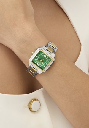 Ručni sat sa zelenim kvadratnim brojčanikom, zlatno-srebrnom metalnom narukvicom na zapešću osobe, pored bijele tkanine sa zlatnim rubom na dugmetu.