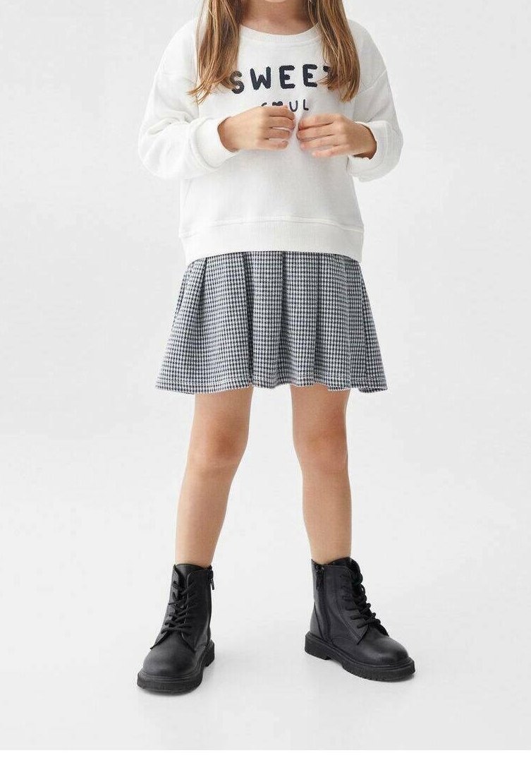 Enfant portant un sweat-shirt blanc avec un texte, une jupe à carreaux noir et blanc, et des bottes noires à lacets, debout devant un fond uni.