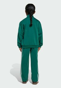 Dijete s dugom pletenicom stoji okrenuto leđima, odjeveno u zeleni trenirka s bijelim prugama i crnim cipelama, na bijeloj pozadini.