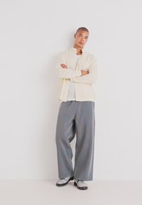 Camisa amarillo claro con botones sobre una blusa gris texturizada, combinada con pantalones anchos grises con costuras azules y zapatos negros.